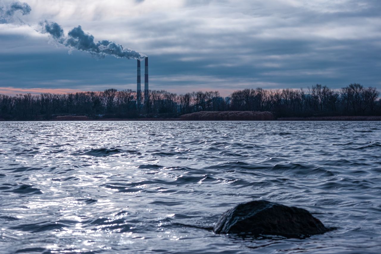 Factory smoke stacks emitting pollution over river with dramatic sky in Kyiv.
